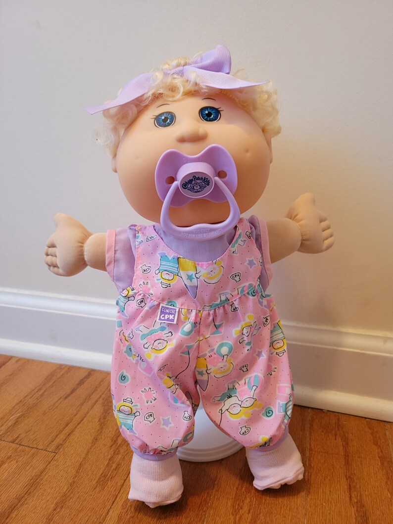 May include: A Cabbage Patch Kids doll with blonde curly hair, blue eyes, and a light purple bow. The doll wears a light purple shirt under pink overalls featuring a colourful pattern of small figures and stars, with a matching light purple pacifier.