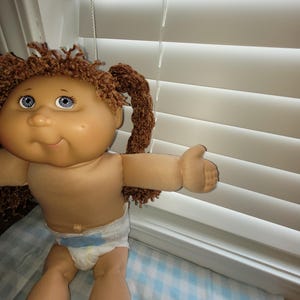 Puede incluir: Una mu&ntilde;eca vintage con una cara redonda, ojos azules y cabello casta&ntilde;o rizado peinado en coletas. La mu&ntilde;eca lleva un pa&ntilde;al y tiene los brazos extendidos. La mu&ntilde;eca est&aacute; sentada sobre una manta a cuadros azules y blancos.