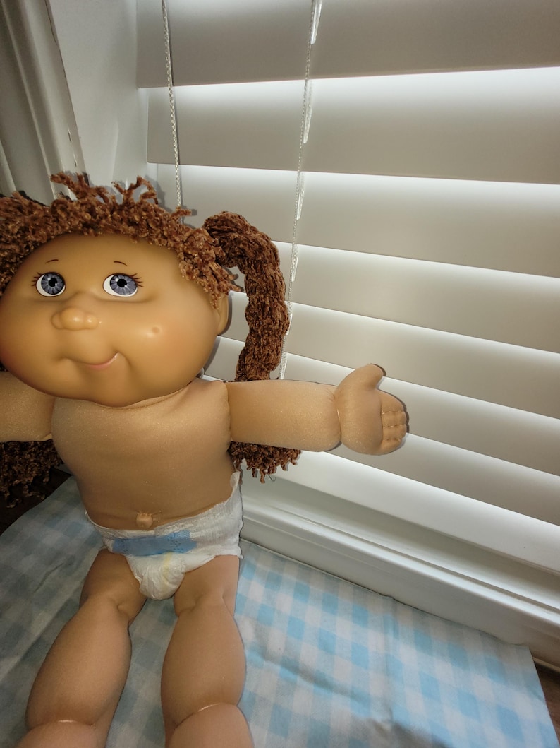 Puede incluir: Una mu&ntilde;eca vintage con una cara redonda, ojos azules y cabello casta&ntilde;o rizado peinado en coletas. La mu&ntilde;eca lleva un pa&ntilde;al y tiene los brazos extendidos. La mu&ntilde;eca est&aacute; sentada sobre una manta a cuadros azules y blancos.
