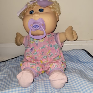 May include: A Cabbage Patch Kids doll with blonde curly hair, blue eyes, and a light purple bow. The doll wears a pink romper with a multi-coloured pattern and a purple pacifier in its mouth. The doll has light skin and is wearing light pink socks.