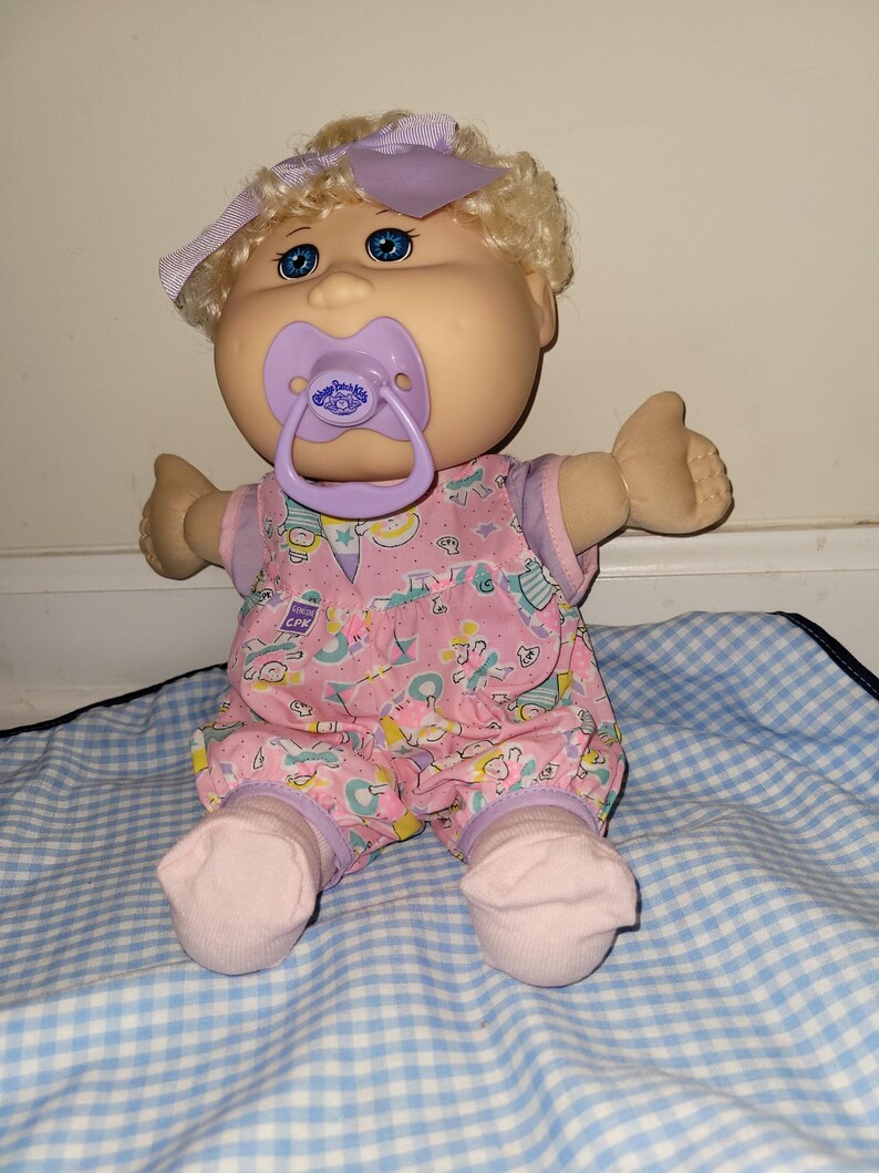 May include: A Cabbage Patch Kids doll with blonde curly hair, blue eyes, and a light purple bow. The doll wears a pink romper with a multi-coloured pattern and a purple pacifier in its mouth. The doll has light skin and is wearing light pink socks.