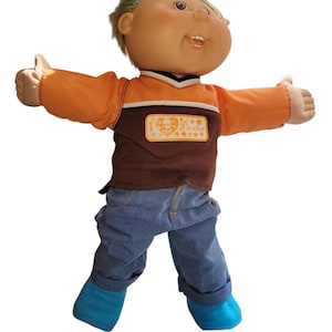 May include: A vintage doll with a smiling face, wearing an orange and brown shirt with a pirate-themed patch, blue jeans, and blue boots. The doll has green hair and is posed with arms outstretched.