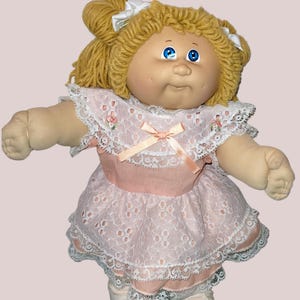 May include: A vintage doll with blonde yarn hair styled in pigtails, adorned with white bows. The doll wears a pink dress with lace trim and a matching pink bow. The doll has blue eyes and is wearing white shoes.