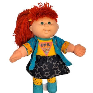 May include: A doll with bright orange yarn hair, green eyes, and freckles. It wears a yellow shirt with a pink triangle patch reading CPK Kids, a teal open jacket with floral cuffs, a black skirt with white star patterns, yellow polka-dot leggings, and teal shoes.
