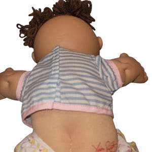 May include: A doll with brown yarn hair wearing a light blue and white striped shirt with pink trim and patterned trousers. The doll has a red signature on its lower back. The doll is viewed from the back, showing the shirt closure and hair texture.