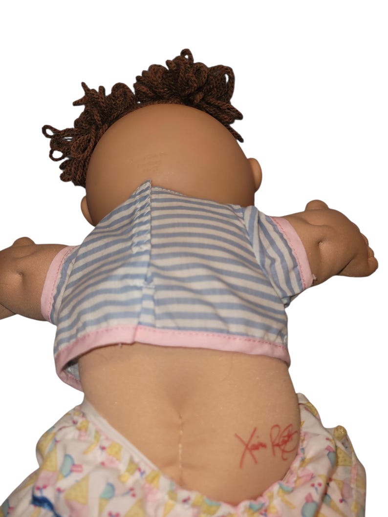 May include: A doll with brown yarn hair wearing a light blue and white striped shirt with pink trim and patterned trousers. The doll has a red signature on its lower back. The doll is viewed from the back, showing the shirt closure and hair texture.