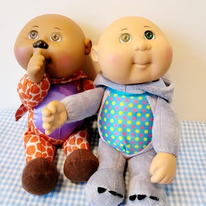 May include: Two Cabbage Patch Kids dolls. One doll wears a giraffe-patterned outfit with a purple torso. The other doll is in a gray onesie with a blue and yellow polka dot pattern. Both dolls have bald heads and painted facial features.