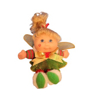 May include: A doll with blonde hair tied in a ponytail, blue eyes, and a red flower mark on its cheek. It wears a green and red striped long-sleeve top, a metallic green skirt with a gold ribbon bow, silver wings, and green shoes with white fur trim.