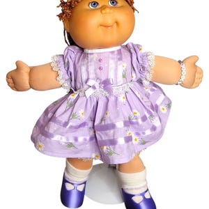 May include: A soft-sculpture doll with brown yarn hair, blue eyes, and a light skin tone. The doll wears a lavender dress with a white daisy print, lace trim, and a small bow, paired with white socks and shiny purple shoes. A beaded bracelet is on one wrist.
