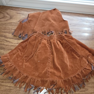 Op de afbeelding: Een bruine su&egrave;de rok en vest set. De rok heeft een gefranjerde zoom. Het vest is mouwloos met franjes langs de onderkant. De set is gemaakt van een zacht, su&egrave;de-achtig materiaal.