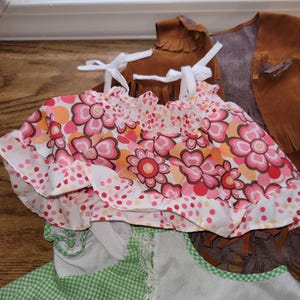 Op de afbeelding: Een verzameling kinderkleding, waaronder een top met bloemenprint met strikbandjes en ruches, een groen geruit jurkje met kanten rand en een bruin su&egrave;de vest met franjes.