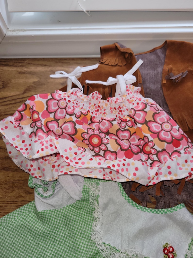 Op de afbeelding: Een verzameling kinderkleding, waaronder een top met bloemenprint met strikbandjes en ruches, een groen geruit jurkje met kanten rand en een bruin su&egrave;de vest met franjes.