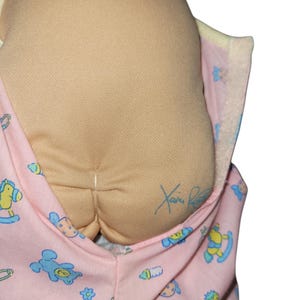 May include: A soft fabric doll with a tan, rounded head featuring a signature in blue ink. The doll is wrapped in a light pink swaddle blanket decorated with a repeating pattern of blue teddy bears, yellow rocking horses, and green strawberries.