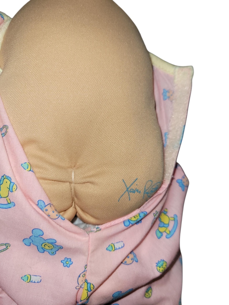 May include: A soft fabric doll with a tan, rounded head featuring a signature in blue ink. The doll is wrapped in a light pink swaddle blanket decorated with a repeating pattern of blue teddy bears, yellow rocking horses, and green strawberries.