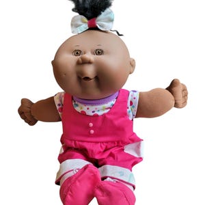 May include: A vintage Cabbage Patch Kid doll with dark skin, black hair, and brown eyes. The doll wears a pink outfit with a white and colorful polka dot shirt underneath. A bow adorns the top of the doll's head.