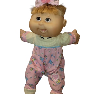 May include: A doll with blonde hair tied in a ponytail, featuring a pink butterfly hair accessory. The doll wears a cream-coloured top and pink trousers with a pattern of small blue bears and toys, including a heart-shaped patch with the text CPK.