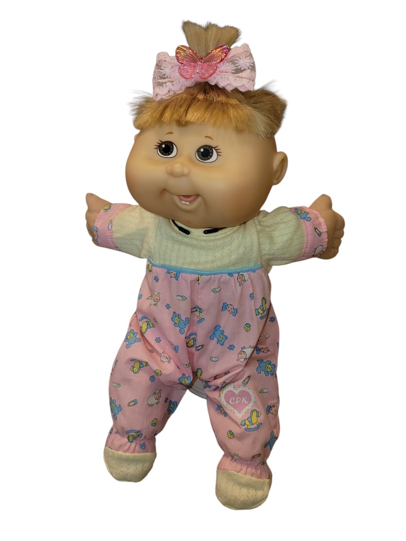 May include: A doll with blonde hair tied in a ponytail, featuring a pink butterfly hair accessory. The doll wears a cream-coloured top and pink trousers with a pattern of small blue bears and toys, including a heart-shaped patch with the text CPK.