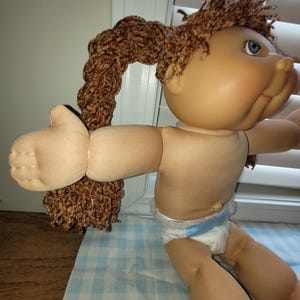 Puede incluir: Una mu&ntilde;eca vintage con tez bronceada, ojos azules y cabello casta&ntilde;o peinado en dos coletas. La mu&ntilde;eca lleva un pa&ntilde;al. Los brazos de la mu&ntilde;eca est&aacute;n extendidos.