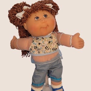 May include: A vintage doll with brown yarn hair styled in pigtails, adorned with white bows. The doll wears a floral top, denim shorts, and blue shoes. A bracelet is on the doll's wrist. The doll is standing on a white stand.