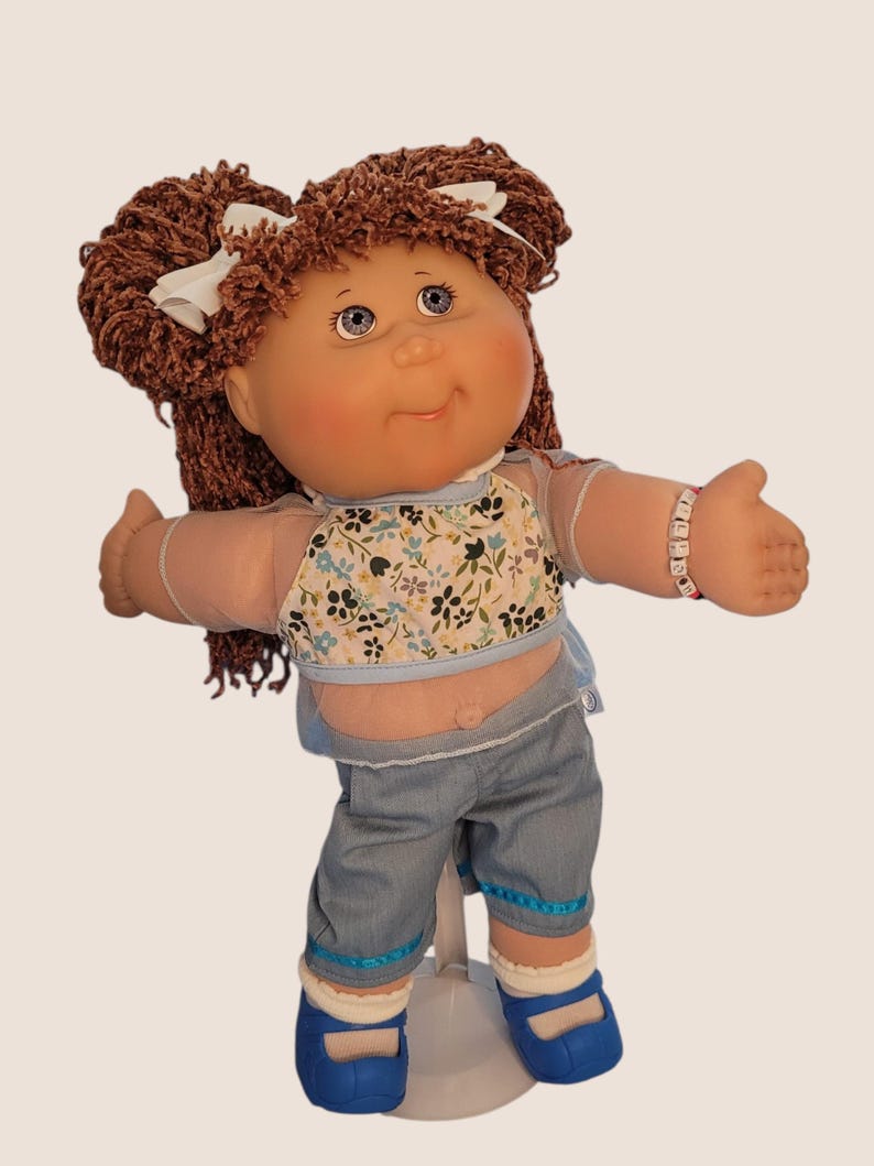 Puede incluir: Una mu&ntilde;eca vintage con cabello casta&ntilde;o de hilo en coletas, adornadas con lazos blancos. La mu&ntilde;eca viste una blusa floral, pantalones cortos de mezclilla y zapatos azules. Una pulsera est&aacute; en la mu&ntilde;eca de la mu&ntilde;eca.