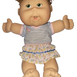May include: A Cabbage Patch Kids doll with brown yarn hair, green eyes, and a light skin tone. The doll wears a blue and white striped short-sleeve shirt with a pink trim and a white skirt featuring a colourful ice cream cone pattern.