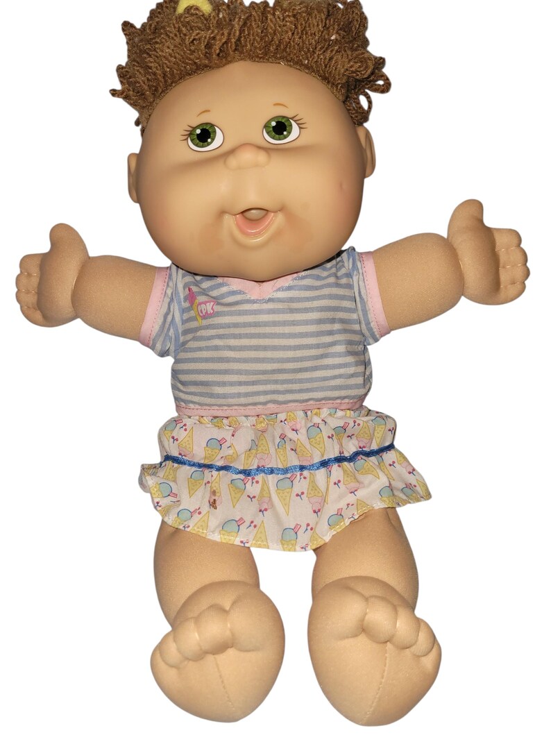 May include: A Cabbage Patch Kids doll with brown yarn hair, green eyes, and a light skin tone. The doll wears a blue and white striped short-sleeve shirt with a pink trim and a white skirt featuring a colourful ice cream cone pattern.