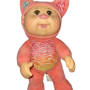 May include: A Cabbage Patch Kids doll wearing a pink cat costume. The doll has a round face, brown eyes, and a pink nose. The costume features a hood with cat ears and a zebra-striped design on the chest. The doll's arms and legs are a light tan color.