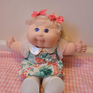 May include: A vintage Cabbage Patch Kid doll with blonde hair and blue eyes. The doll wears a floral dress with a green and white checkered bodice, white shoes, and pink bows. The doll is sitting on a pink and white checkered surface.