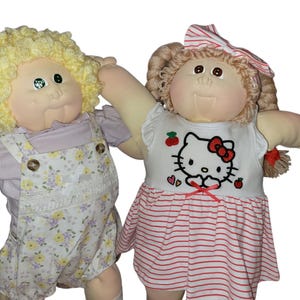 May include: Two dolls with soft fabric bodies and yarn hair. The left doll has curly yellow hair and wears lavender overalls with a floral print. The right doll has light brown braided hair with a striped bow and a white dress featuring Hello Kitty.