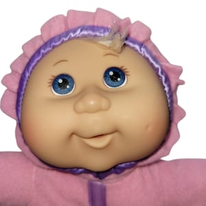 May include: A vintage doll with a soft, vinyl face and blue eyes. The doll is wearing a pink hooded outfit with a purple trim. The doll has a small tuft of blonde hair and rosy cheeks. The doll's face has a gentle, smiling expression.