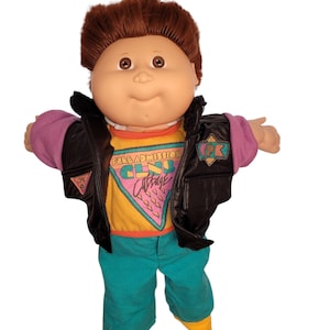 May include: A doll with brown hair wearing a black vest over a yellow shirt with the text Free Admission Club Cabbage, purple sleeves, and teal pants. The doll has brown eyes and a soft-sculpted face with a small nose and a closed mouth.