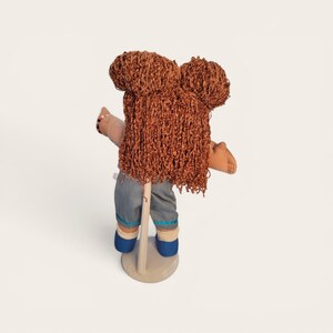 Puede incluir: Mu&ntilde;eca con cabello de hilo marr&oacute;n peinado en dos mo&ntilde;os y flequillo largo. La mu&ntilde;eca lleva vaqueros azules con ribetes azules y beige, y zapatos azules. La mu&ntilde;eca est&aacute; montada sobre un soporte beige.