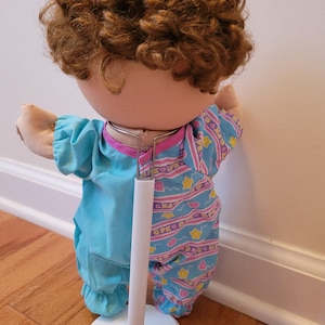 May include: A doll with curly brown hair wearing a two-toned outfit. The left side is solid light blue, while the right side features a light blue pattern with pink squiggly lines, yellow stars, and the text CPK. The doll is supported by a white stand.