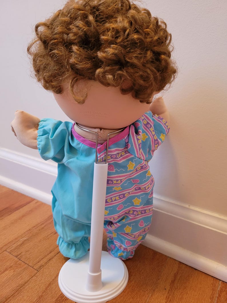 May include: A doll with curly brown hair wearing a two-toned outfit. The left side is solid light blue, while the right side features a light blue pattern with pink squiggly lines, yellow stars, and the text CPK. The doll is supported by a white stand.