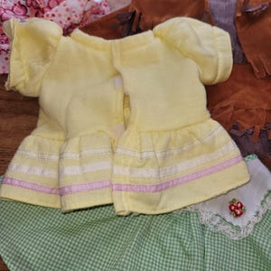 Op de afbeelding: Een verzameling poppenkleding, waaronder twee gele jurken met gefranjerde mouwen en rokken, een groen-witte geruite rok en een bruin vest met franjes. Andere items zijn een roze bloemenjurk en bruine laarzen.