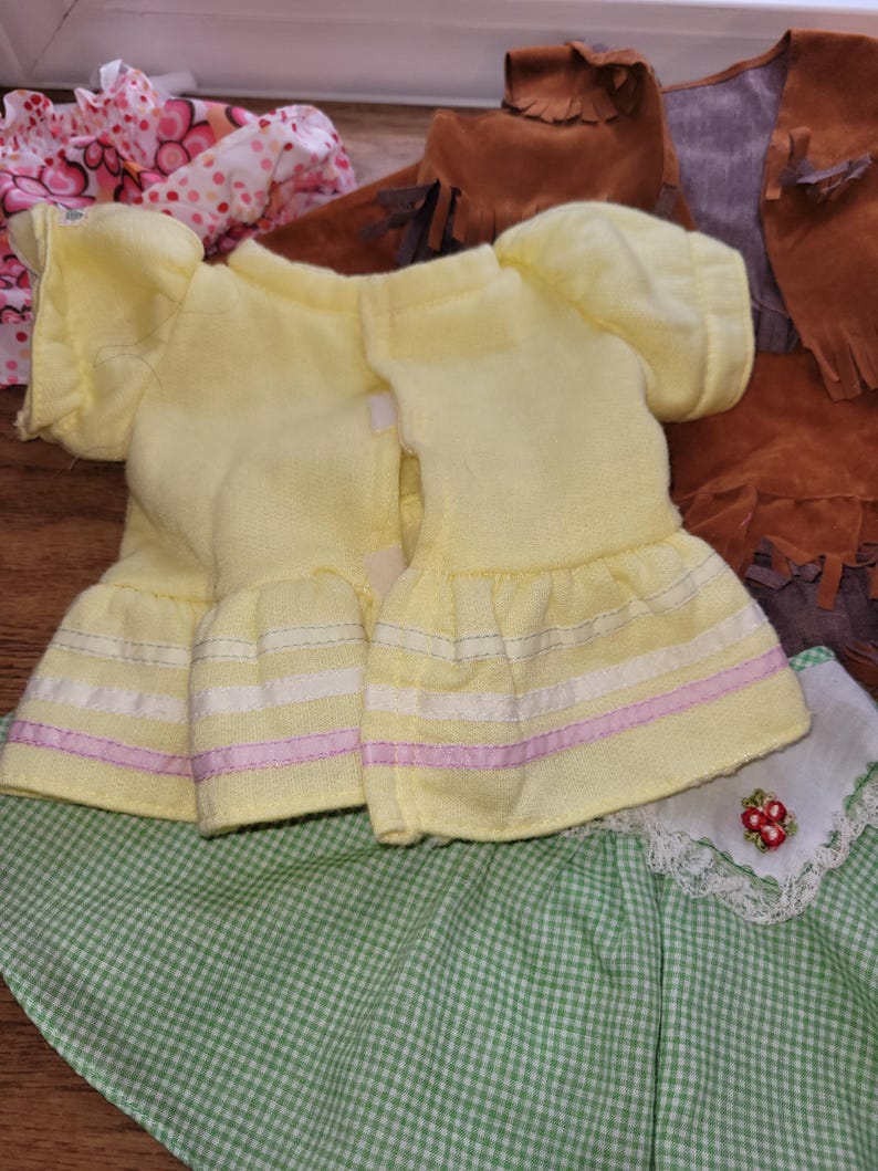 Op de afbeelding: Een verzameling poppenkleding, waaronder twee gele jurken met gefranjerde mouwen en rokken, een groen-witte geruite rok en een bruin vest met franjes. Andere items zijn een roze bloemenjurk en bruine laarzen.