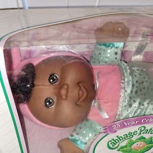 May include: A Cabbage Patch Kids doll in its original packaging, featuring a 25 Year Celebration label. The doll has dark skin, black hair, and brown eyes, wearing a pink top with a white ribbon and light green pants with a silver star pattern.