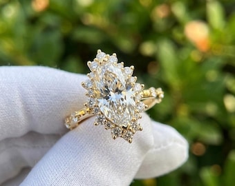 May include: A gold engagement ring with a large, marquise-cut diamond centre stone, encircled by a halo of smaller diamonds. The band also features small diamonds. The ring is set against a blurred green background.