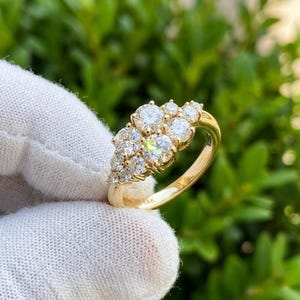 May include: A gold ring featuring a cluster of round, clear gemstones. The ring has a polished gold band and is marked with "14K". The gemstones are arranged in a floral or cluster design, creating a sparkling effect.