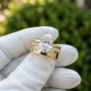 May include: A set of three gold rings. The engagement ring features a large, oval-cut diamond. The wedding band is adorned with small diamonds, and the third ring has a clover design. The rings are held against a blurred green background.