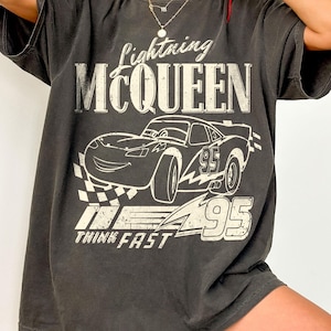 May include: Dark gray t-shirt featuring Lightning McQueen from the movie Cars. The graphic includes the text "Lightning McQueen", a drawing of the race car, the number "95", and the words "Think Fast".