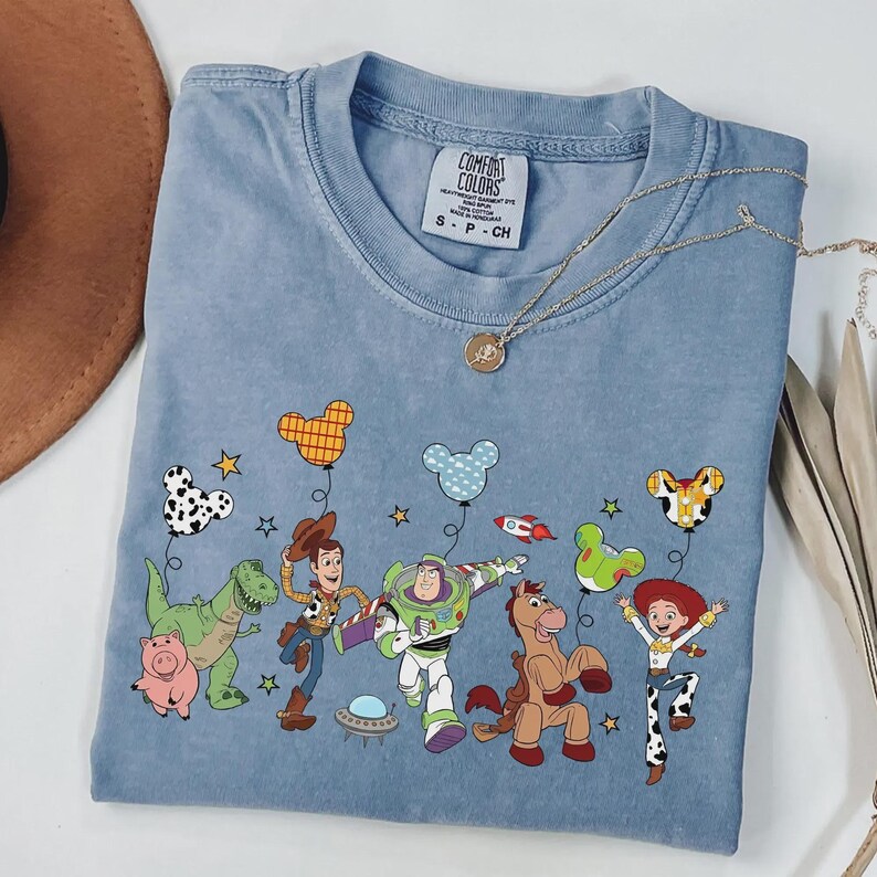 Może przedstawiać: Jasnoniebieska koszulka Comfort Colors z kolorową grafiką postaci z Toy Story. Projekt zawiera Woody'ego, Buzz'a Astrala, Jessie i inne postacie z balonami w kształcie Myszki Miki. Złoty naszyjnik jest zarzucony na koszulkę.