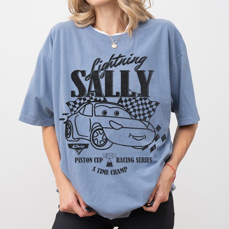 May include: A blue t-shirt featuring a graphic of a cartoon car with the text "Lightning Sally". The design includes a checkered flag and the words "Piston Cup Racing Series" and "5 Time Champ".