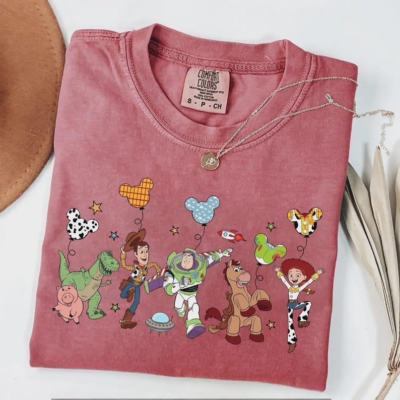 Może przedstawiać: Koszulka Comfort Colors w kolorze przydymionego r&oacute;żu z grafiką postaci z Toy Story i balonami. Postacie to m.in. Woody, Buzz Lightyear i Jessie. Widoczny jest r&oacute;wnież srebrny naszyjnik z wisiorkiem.