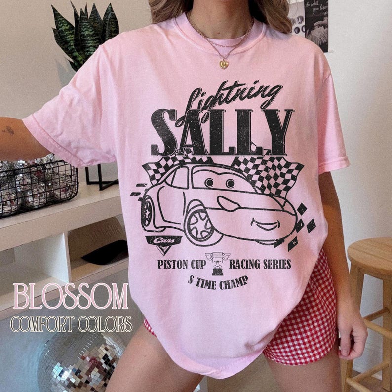 May include: A light pink t-shirt with a black graphic of a cartoon car and the words "Lightning Sally." The shirt also features the text "Piston Cup Racing Series" and "5 Time Champ."