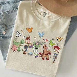 Może przedstawiać: Kremowa koszulka z kolorową grafiką postaci z Toy Story z balonami w kształcie Myszki Miki. Projekt zawiera Woody'ego, Buzz'a Astrala i inne postacie. Koszulka jest złożona na niebieskich dżinsach.