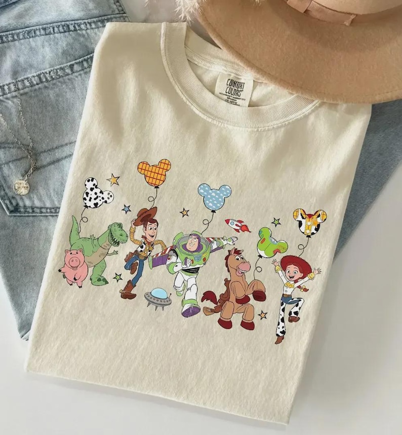 Może przedstawiać: Kremowa koszulka z kolorową grafiką postaci z Toy Story z balonami w kształcie Myszki Miki. Projekt zawiera Woody'ego, Buzz'a Astrala i inne postacie. Koszulka jest złożona na niebieskich dżinsach.