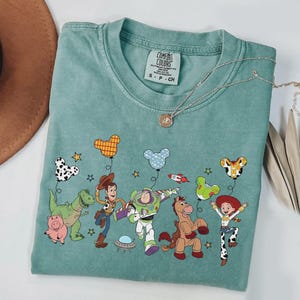 Może przedstawiać: Miętowa koszulka Comfort Colors z grafiką postaci z Toy Story. Projekt zawiera Woody'ego, Buzza Astrala, Jessie i inne postacie z balonami. Widoczny jest r&oacute;wnież srebrny naszyjnik z wisiorkiem.