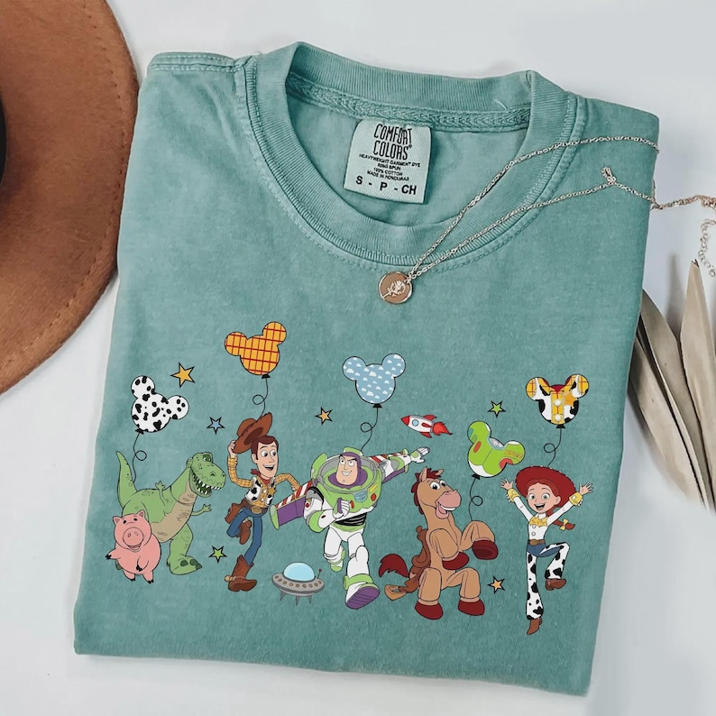 Może przedstawiać: Miętowa koszulka Comfort Colors z grafiką postaci z Toy Story. Projekt zawiera Woody'ego, Buzza Astrala, Jessie i inne postacie z balonami. Widoczny jest r&oacute;wnież srebrny naszyjnik z wisiorkiem.