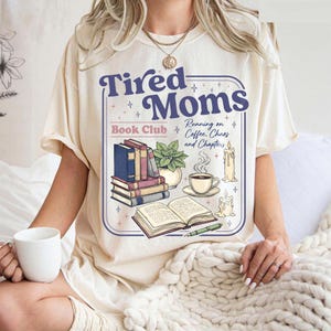 Può includere: T-shirt color crema con la scritta "Tired Moms Book Club". Il design include pile di libri, una tazza di caffè, candele e una pianta. La maglietta ha maniche corte e una vestibilità ampia.